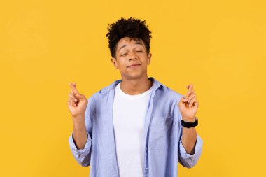 İyimser genç siyah erkek öğrenci gözleri kapalı, şans dilemek için şans diliyor, parlak sarı arka planda hüsnükuruntu yapıyor.