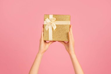 Eller zarif bir şekilde altın renkli bir hediye kutusunu zarif bir krem kurdeleyle sunuyor. Yumuşak pembe bir arka planda hediye vermeyi, kutlamayı ve sürprizi sembolize ediyor.