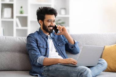 Gözlüklü mutlu Hintli adam evde dizüstü bilgisayar kullanırken telefonla konuşuyor. Genç doğulu serbest çalışan erkek oturma odasındaki koltukta rahatça oturuyor ve uzaktan çalışma verimliliği gösteriyor.