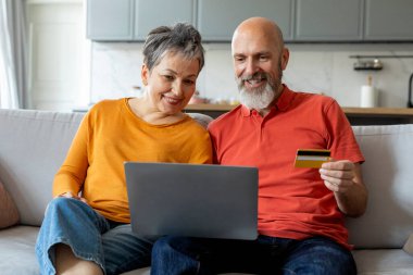 Evde online alışveriş için dizüstü bilgisayar ve kredi kartı kullanan yaşlı çiftler, modern teknolojilerin tadını çıkaran yaşlı eşler, internet alışverişi yapan mutlu erkek ve kadın, yakın çekim