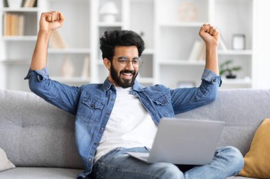 Kot gömlekli neşeli Hintli adam dizüstü bilgisayar ekranına bakarken yumruklarını kaldırıyor, rahat oturma odasında kanepede oturan mutlu doğu çocuğu, başarıyı ve kişisel başarıyı kutluyor.