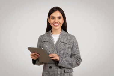 Houndstooth blazer ceket giyen, tablet tutan ve kullanan, kendine güvenen genç iş kadını, gri zemin karşı, hoş bir gülümseme ile işine meşgul