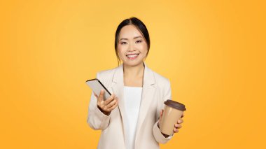 Milenyum çağından kalma takım elbiseli, elinde kahve, akıllı telefonla sohbet eden, sarı stüdyo arka planında izole edilmiş, panorama olan Çinli bir kadın. İş uygulaması, reklam ve teklif, yaşam tarzı