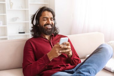 Neşeli sakallı bin yıllık Hintli adam evde telefon ve kulaklık kullanıyor, koltukta oturuyor, video içeriği izliyor veya mobil uygulama üzerinden favori müzik dinliyor, uzayı kopyalıyor.