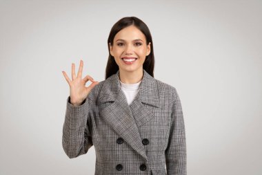 Şık ekose ceketli, kendine güvenen genç bir kadın sağ eliyle imzala açık gri arka planda hoş bir şekilde gülümse.