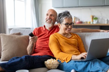 Mutlu, yaşlı bir çift evdeki kanepede birlikte vakit geçiriyor, neşeli eşler oturma odasında dinleniyor, gülümsüyor, televizyon seyrediyor ve uzaktan kumandalı, yaşlı bir kadın dizüstü bilgisayar kullanıyor.