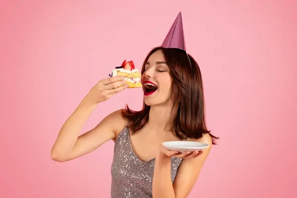 スパークリングシルバーのドレスとピンクのパーティーの帽子で茶色の髪を持つ楽しい女性は イチゴでトッピングされたケーキから噛もうとしています 予想で閉じた目 ピンクの背景に — ストック写真