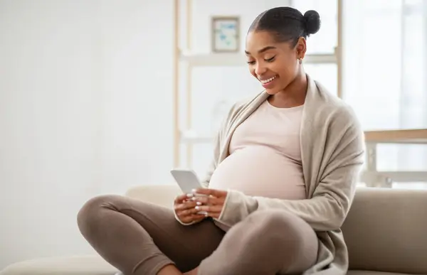 スマートフォンで面白いビデオを見ている美しい若い妊婦は 自宅でソファーに座っている間 モバイルを使用しています 携帯電話 モバイルアプリ コピースペースを使用してアフリカ系アメリカ人女性を期待してミレニアル世代 — ストック写真