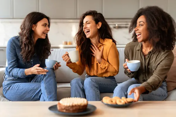 Three joyful multicultural women friends share a cozy moment over coffee and cake, celebrating their friendship in the modern kitchen. Warm moments of true female friendship