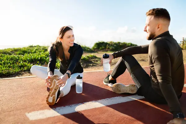 Enthusiastic young couple in workout attire enjoy conversation and laugh during a stretching session on a bright, sunny running track by the sea