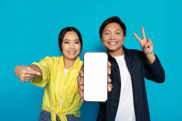 stock image A cheerful Asian couple takes a selfie with a smartphone, the woman pointing at the screen and the man showing a peace sign, both with big smiles against a turquoise background