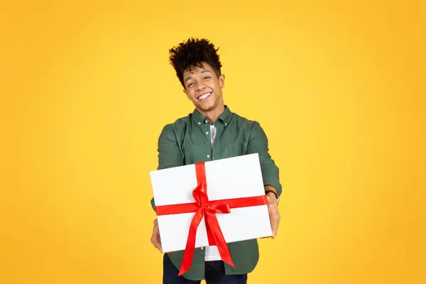 快乐英俊的黑人小伙子送给礼物的白色礼品盒 上面有红色的蝴蝶结 对着相机微笑 在黄色的背景下显得孤立无援 生日聚会 情人节庆祝活动 — 图库照片