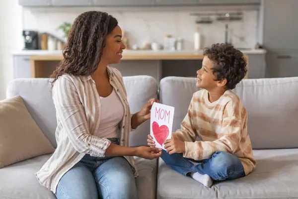 Mothers Day. Little Son Giving Greeting Card To His Happy Mom At Home, Loving Preteen Male Child Congratulating Mommy With Holiday While Sitting Together On Couch In Living Room, Free Space