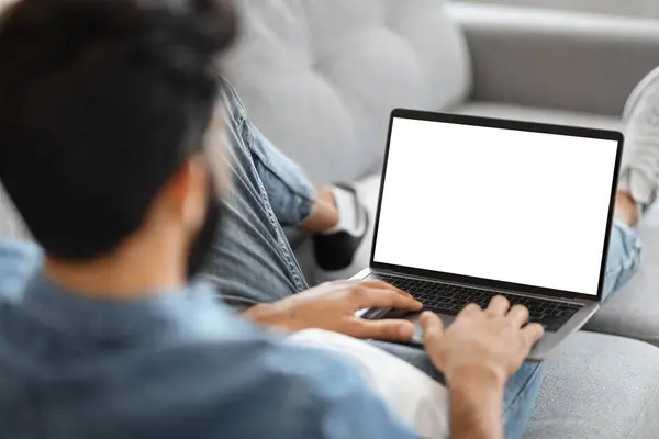 Indian Man Using Laptop Computer With Blank White Screen While Sitting On Couch At Home, Unrecognizable Male Working Online Or Browsing Internet Website, Over Shoulder View, Mockup With Copy Space