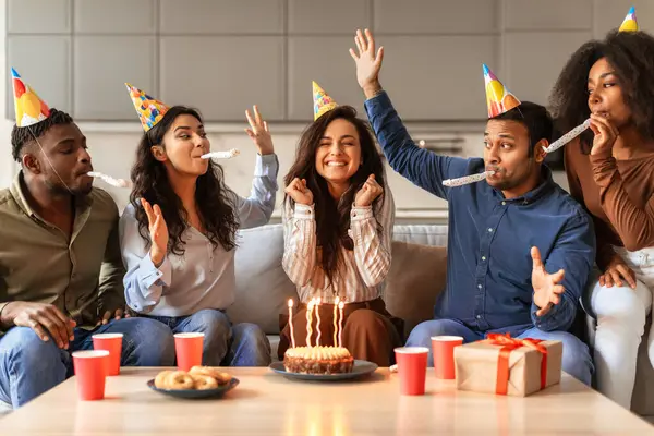 Diverse ladies and guys in festive hats gathering to make birthday wish near cake, their celebration filled with fun and true friendship at home party around table. Woman enjoying bday with friends