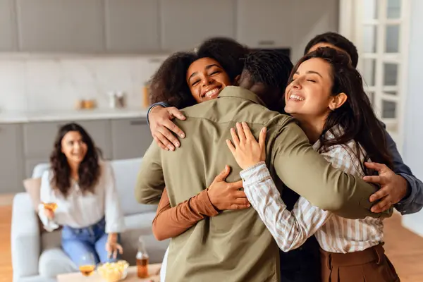 Group of diverse young friends sharing hug during home party gathering at modern apartment interior. Fellow students, men and women, embracing united in laughter and friendship on weekend