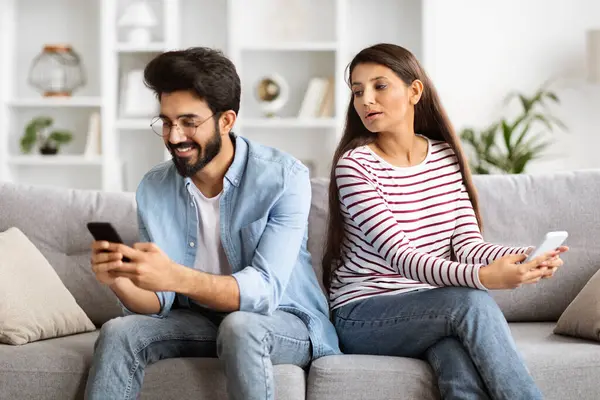 Shocked indian woman spying on her smiling boyfriend who using smartphone, chatting or scrolling social media news feed, chatting with girls. Couple sitting on couch at home, using phones