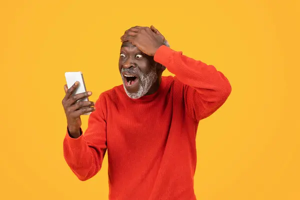 兴奋地震惊着身穿红衣的年长黑人男子 张开嘴 看着智能手机 隔离在橙色工作室的背景下 好消息 有趣的视频 应用程序和设备 — 图库照片