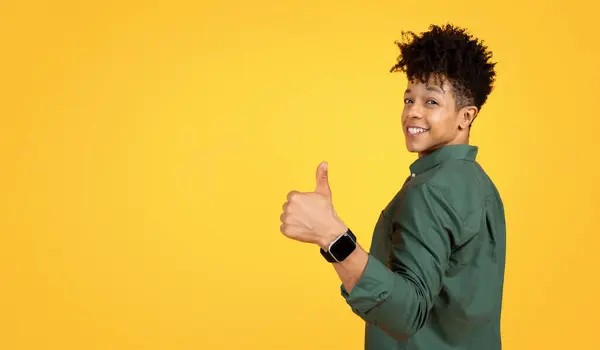 年轻帅气的年轻黑人小伙子穿着绿色的衬衫 对着摄像机露出大拇指和微笑 被黄色的摄影棚背景隔离 还有广告的复制空间 — 图库照片