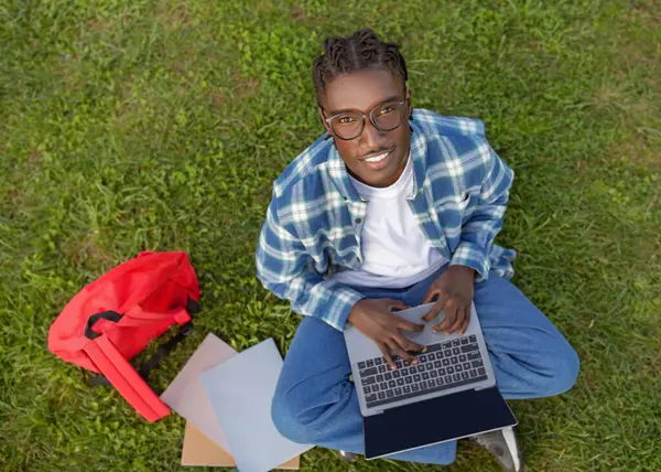 Black college student guy typing on laptop PC while sits outside on green grass in park, engaged in distance online learning leisure, smiling to camera, above view shot