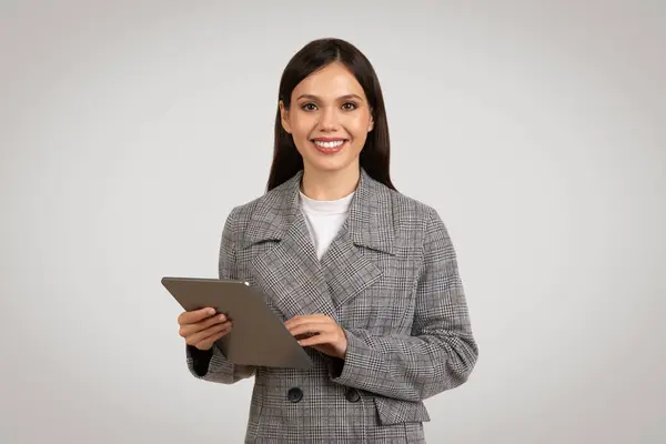 自信を持った若いビジネスウーマン ハウンドスブレザー 保持し タブレットを使用して 快適な笑顔で彼女の仕事に従事 グレーの背景に対して — ストック写真