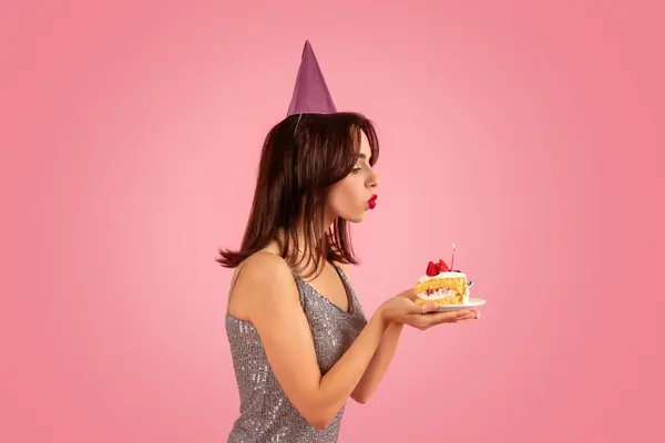 明るいドレスとパーティーハットを着たパーティーの気分で陽気な女性は 優しくバースデーケーキのピンクの背景にろうそくを吹き飛ばします ホリデーイベント — ストック写真