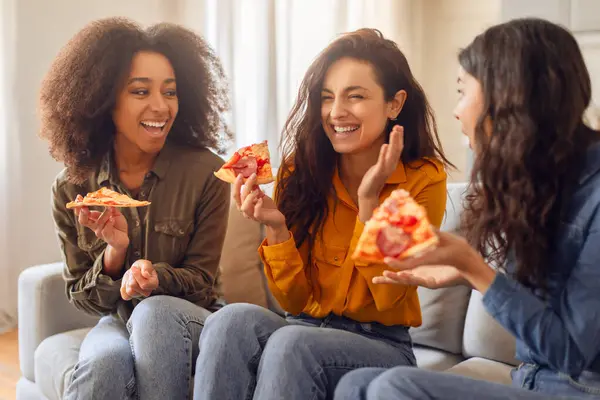 Three diverse women friends enjoying snacks and joyful chat, laughing together while eating pizza on a cozy sofa in modern living room interior. Home gathering, female friendship