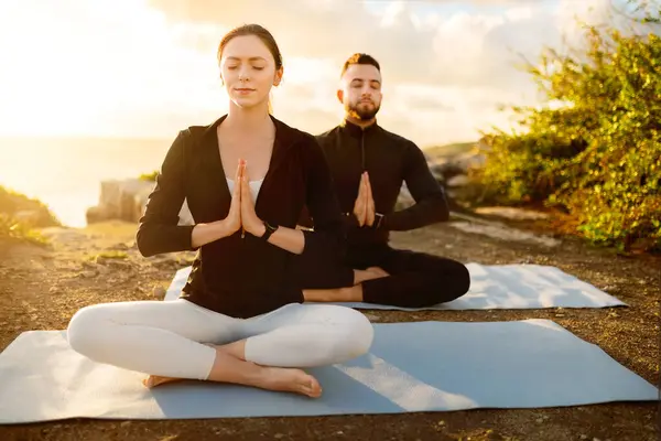 Focused couple in seated meditation pose on yoga mats, enjoying tranquil moment of reflection as the setting sun bathes them in a soft light
