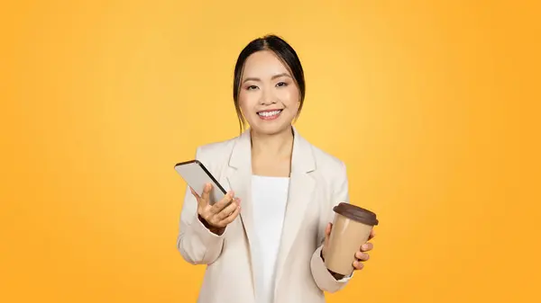 Glad millennial chinese lady in suit with cup of takeaway coffee, chatting on smartphone, isolated on yellow studio background, panorama. Work app, ad and offer, lifestyle