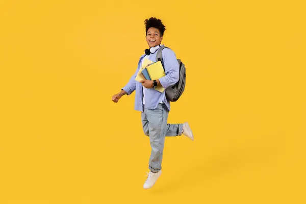 快乐的年轻黑人男生在运动 带着书本和背包跳跃 在明亮的黄色背景下散发着热情和自由 全身而退 — 图库照片