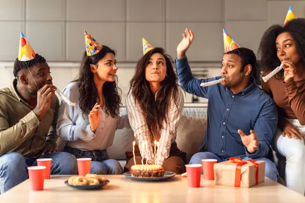 Students enjoying a weekend birthday party, woman in festive hat making wish before blowing out candles and smiling, diverse community coming together in celebration indoors