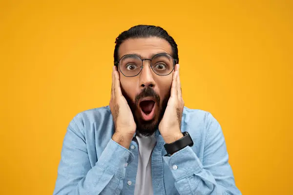 stock image Shocked man in a blue shirt with hands on cheeks and mouth wide open, displaying a surprised and astonished expression against a uniform yellow background. Stress, reaction on news