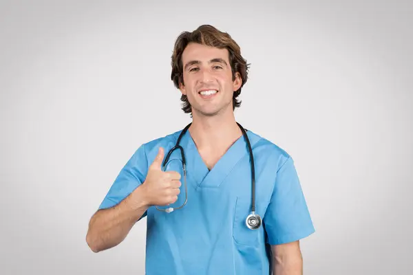 青いスクラブの陽気な若い男性看護師はサインアップし 医療サービス 灰色の背景で成功を示しています ストックフォト