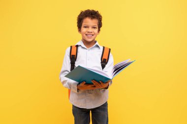 Kitap okumaya dalmış, parlak sarı arka planda kameraya gülümseyen turuncu bir sırt çantası takan zeki siyah bir öğrenci.