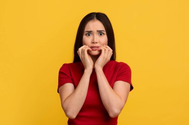 Korkmuş genç Asyalı kadın, büyük korkmuş gözleriyle yüzüne dokunuyor ve kameraya bakıyor, sarı arka planda yalnız poz veriyor. İnsan duyguları kavramı