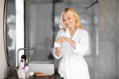 Beyaz bornozlu gülümseyen yaşlı kadın yüz kremi kavanozunu açıyor, mutlu olgun bir kadın modern banyoda cilt bakımına hazırlanıyor, evde güzellik rutininin tadını çıkarıyor, uzay fotokopisini çekiyor.