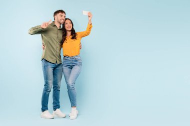 Neşeli genç adam ve kadın gündelik kıyafetlerini giyer selfie çekerken mutlu anları paylaşırlar, barış işareti yapan bir adamla, düz mavi zemine karşı.