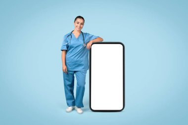 Mavi üniformalı, steteskoplu mutlu kadın hemşire. Büyük bir akıllı telefon ekranına yaslanıyor. Uygulama promosyonu için ideal.
