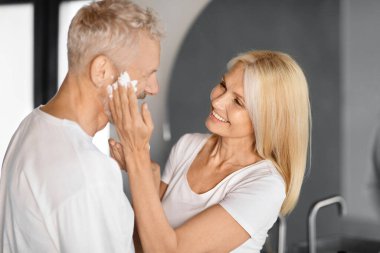 Son sınıf öğrencisi bir kadın kocasına nazikçe nemlendirici krem sürüyor mutlu yaşlı eşler evde modern bir tuvalette cilt bakımını paylaşıyor, yakın plan çekimler yapıyor.