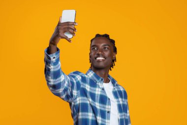 Neşeli genç siyahi adam akıllı telefonuyla selfie çekiyor, mutlu Afrikalı Amerikalı adam telefon kamerasına gülümsüyor, parlak sarı arka planda izole edilmiş poz veriyor, sosyal medya için içerik yaratıyor.