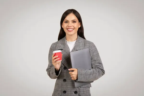 Smiling businesswoman in houndstooth blazer holding red coffee cup and grey laptop, ready for work, against simple light grey background