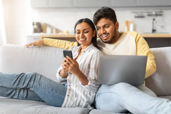 スマートフォンやラップトップとのソファーでリラックスしたインドのカップル 自宅での余暇を楽しんだ幸せな若い東部の配偶者 新しいアプリを閲覧したり ソーシャルネットワークをスクロールしたり 無料のスペース — ストック写真