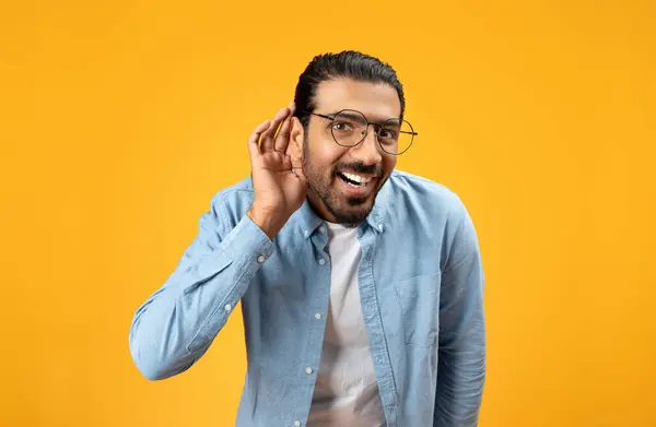 一个穿着蓝色斜纹棉布衬衫的快乐男人 戴着眼镜 用耳朵听着 脸上的表情暗示好奇或偷听 背景是橙色的 — 图库照片