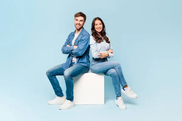 时尚的年轻夫妇穿着斜纹棉布 背靠背地坐着 举止自信 都面带微笑 在浅蓝色的背景下表现出轻松的舒适和冷静的态度 — 图库照片
