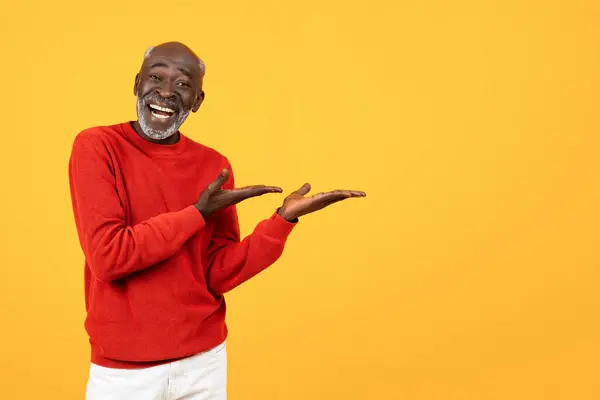 Cheerful elderly Black man in red sweater showcasing with hands on an empty space, against a vivid yellow backdrop, ideal for product placement or advertisement, offer, ad