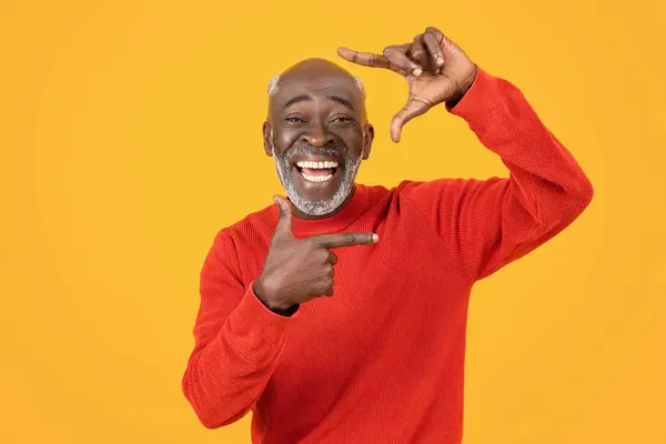 Glad senior black man make frame gesture, hold empty space at hands, isolated on orange studio background. Create art, sale, ad and offer, presenting, facial expression