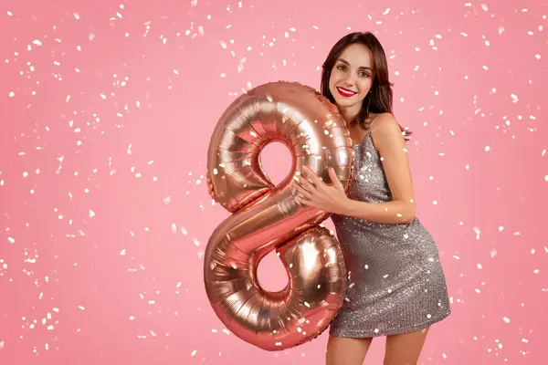 Radiant woman in a shimmering dress holding a rose gold number 8 balloon, surrounded by confetti on a pink background, exuding celebration and joy. Spring holiday celebration event