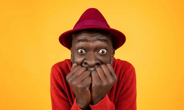 Worried senior Black man in a red sweater and hat, biting his nails with wide eyes full of anxiety against a plain yellow background, showing a humorous expression of suspense