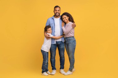 Aile portresi. Mutlu Afrika kökenli Amerikalı ebeveynler ve küçük oğulları sarı arka planda kucaklaşıyorlar, neşeli siyah anne, baba ve erkek çocuk stüdyoda kucaklaşıyorlar.