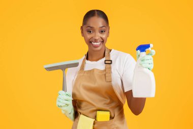 Bahar temizliği. Profesyonel Afro-Amerikan temizleyicisi, cam temizleyicisi ve deterjanıyla, etkin dezenfeksiyon ve hijyeni teşvik ediyor, sarı stüdyo arka planına karşı, ev işlerinden zevk alıyor.